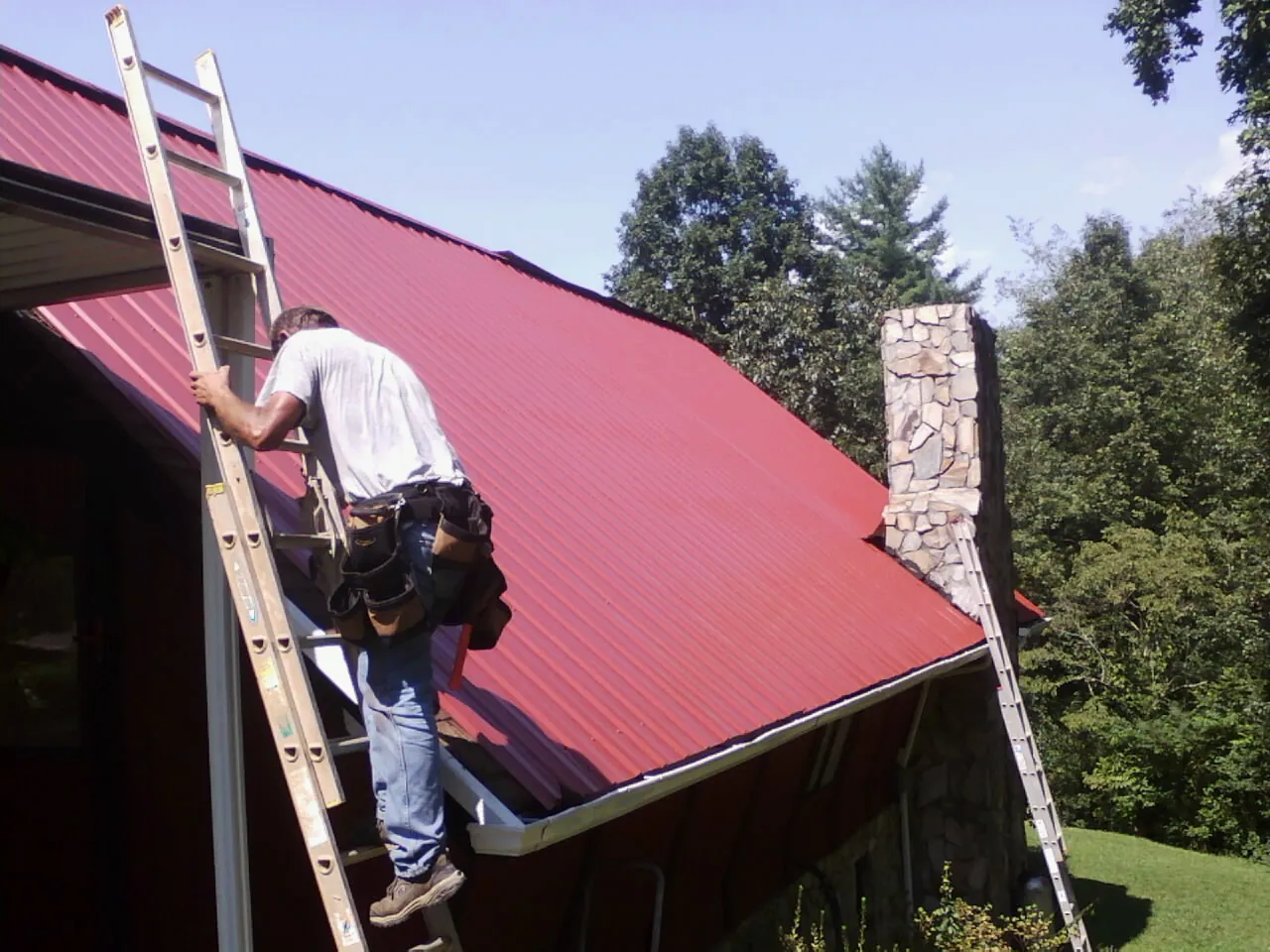 Roofer on a ladder beside a red metal roof and stone chimney — Soffit & Fascia Repair by Roofing Brothers in Pikesville
